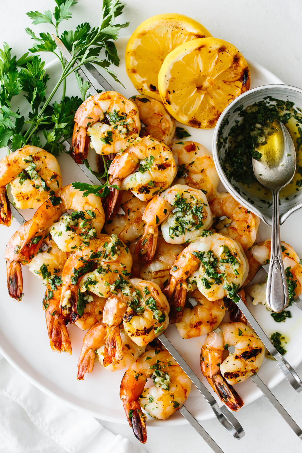 Grilled shrimp skewers on a plate with lemon slices, parsley and the marinade.