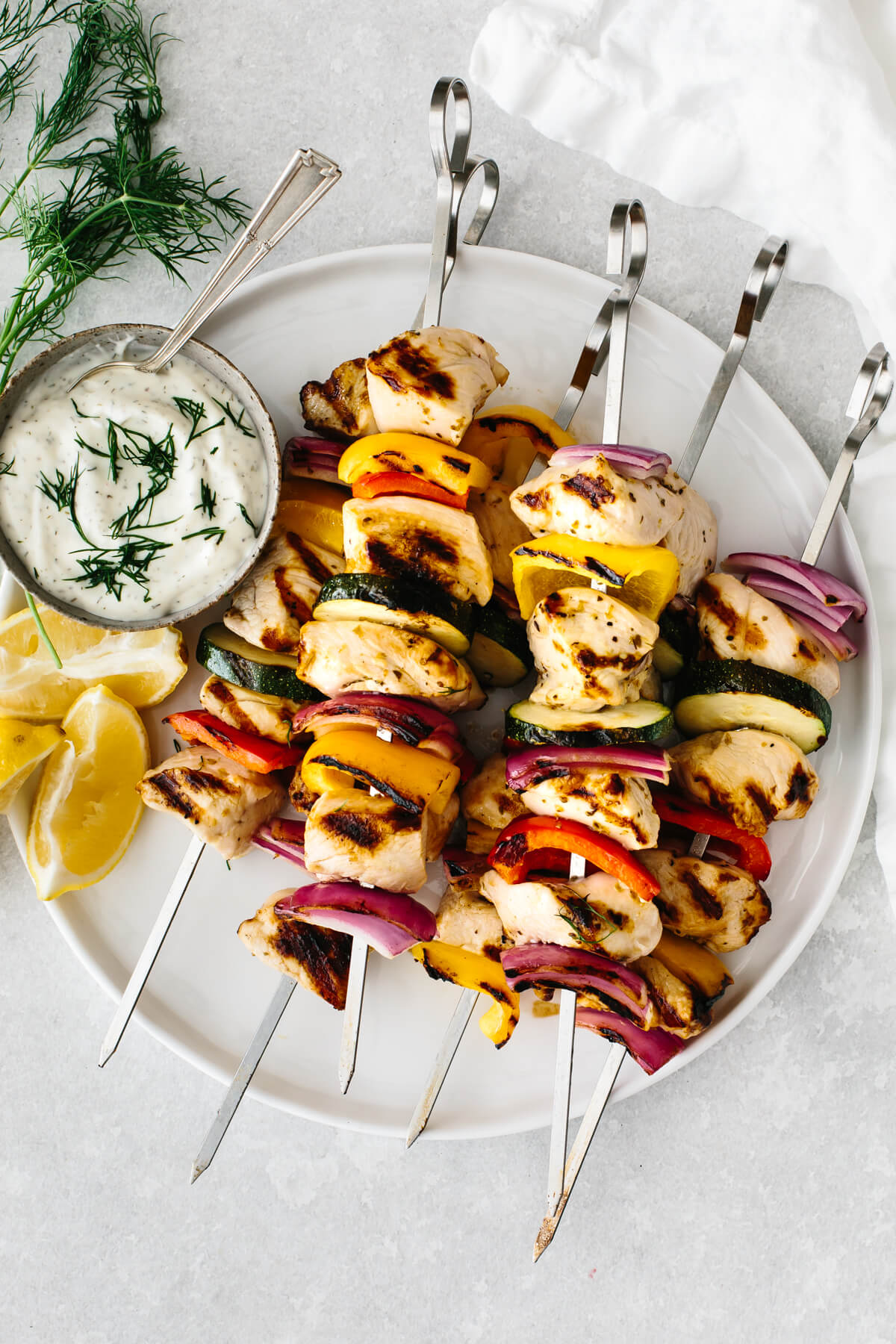 A plate full of cooked chicken kebabs, next to tzatziki sauce.