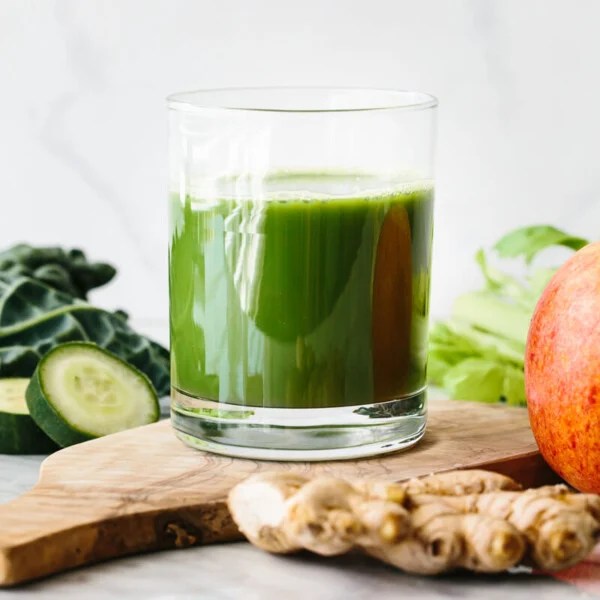 A glass of green juice with an apple, ginger, kale, and cucumber slices around it on a table.
