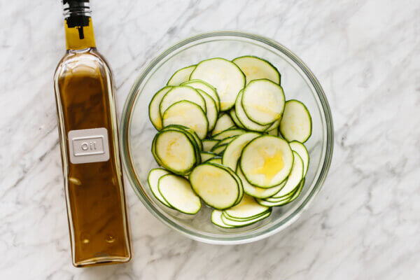 A bowl of fresh zucchini slices next to a glass bottle of olive oil.