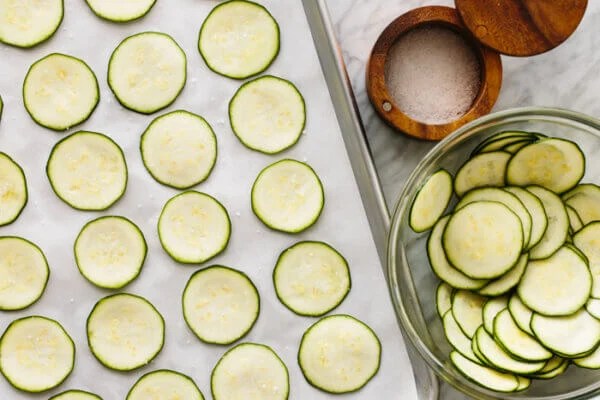 Zucchini slices on top of a parchment lined baking sheet next to a bowl of extra zucchini slices.