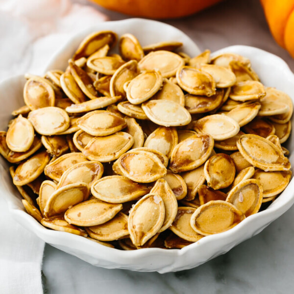 A white bowl with roasted pumpkin seeds next to a pumpkin.