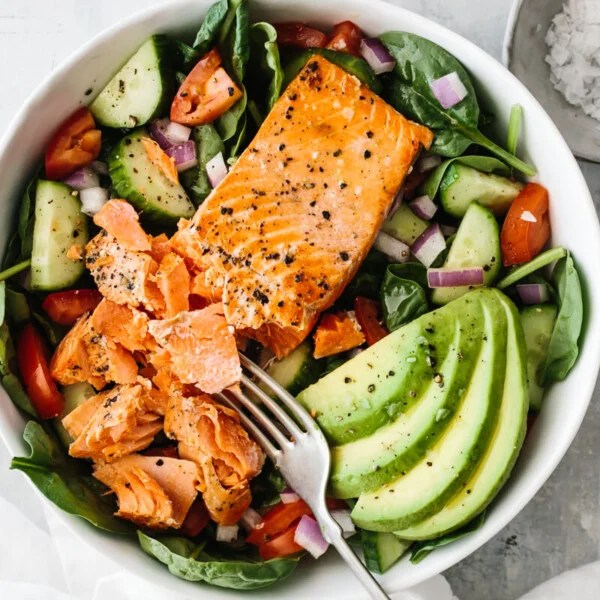 Salmon fillet broken up with fork on top of salad with avocado slices.