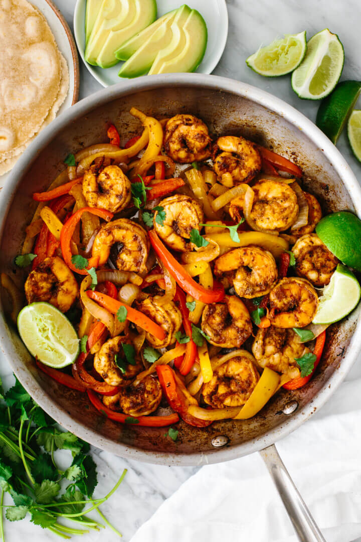 Shrimp fajitas in a pan with avocado slices and lime wedges.