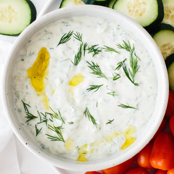 Tzatziki in a bowl topped with a drizzle of olive oil and sprinkle of dill, next to sliced cucumber and cherry tomatoes.