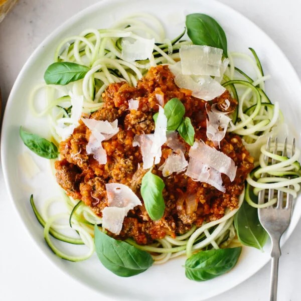 A plate of zucchini noodles topped with bolognese sauce, next to a plate of parmesan.