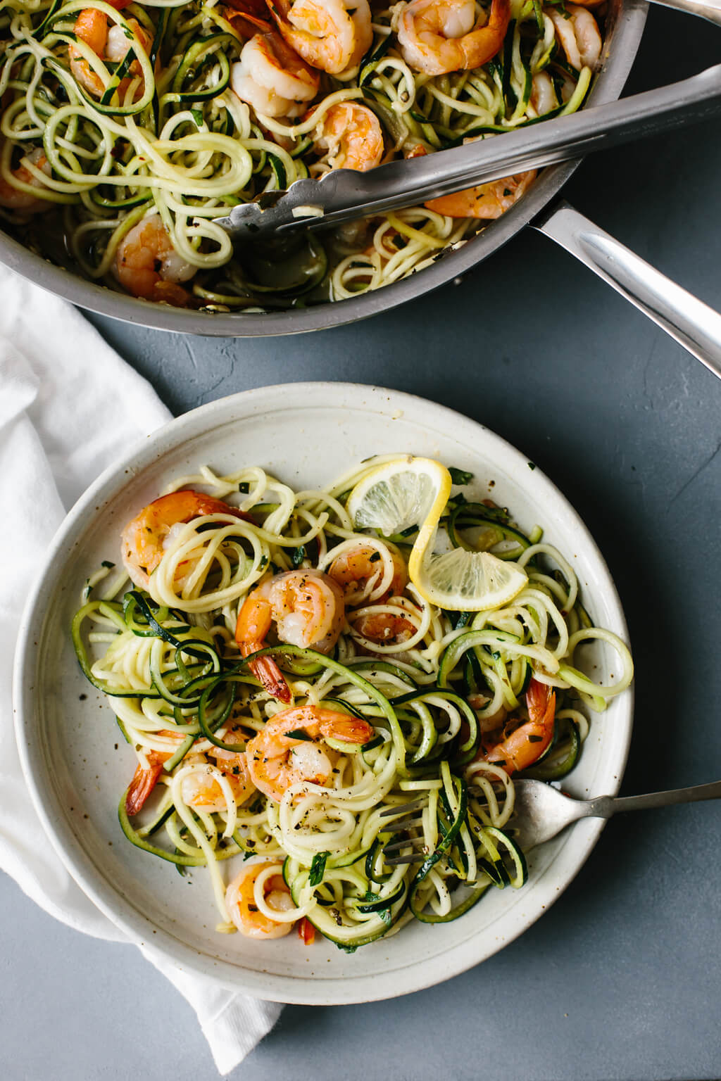 A plate of zucchini pasta noodles and shrimp next to a pan with tongs.