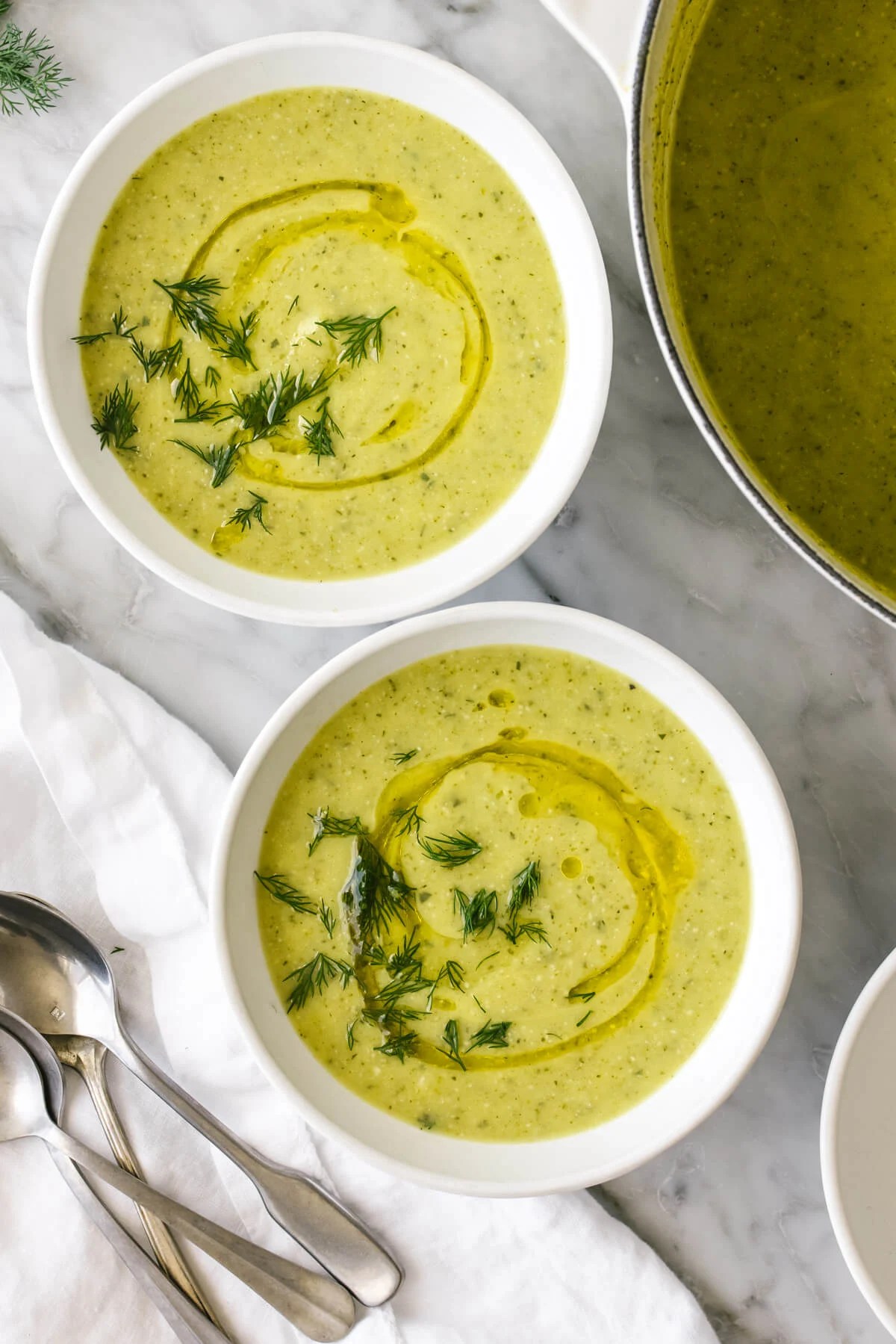 Zucchini soup in two white bowls on a table next to spoons.